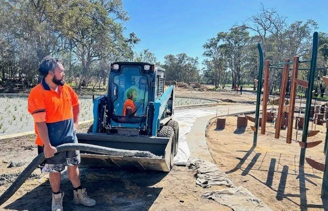 A man in an orange shirt holding a hose, working on landscaping a park with a small construction vehicle nearby, in front of a playground and trees.