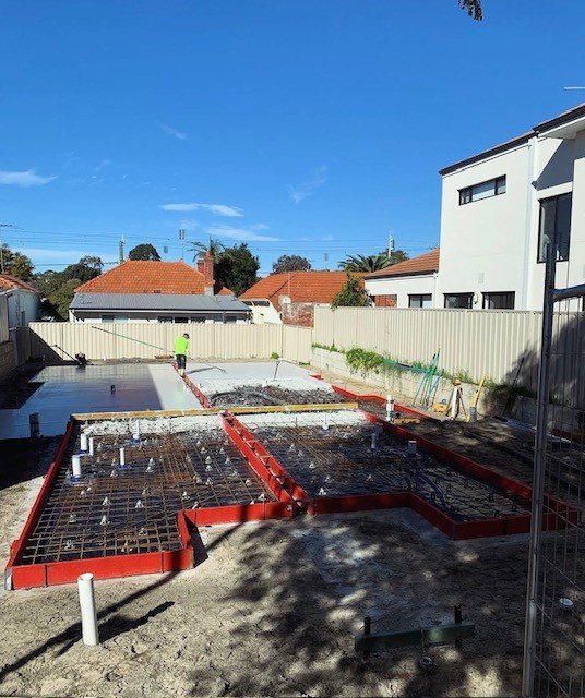 Construction site with red metal framework for a foundation, a worker in a neon shirt working, and neighboring houses under a blue sky.