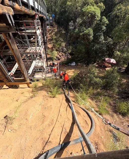 Workers in orange safety gear and helmets setting up a large hose or cable on the ground near a construction or industrial site with a tall metallic structure and trees in the background.