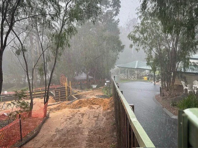 A path and sidewalk separated by a wooden railing during rainy weather, with muddy ground on the left and wet pavement on the right, surrounded by trees and outdoor furniture.