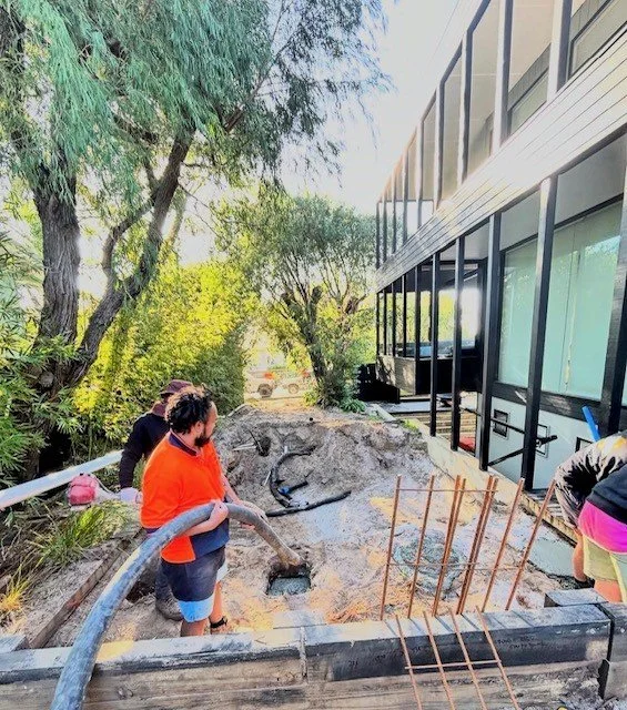 People working on a construction site next to a modern building, with construction pipes and rebar, and trees in the background.