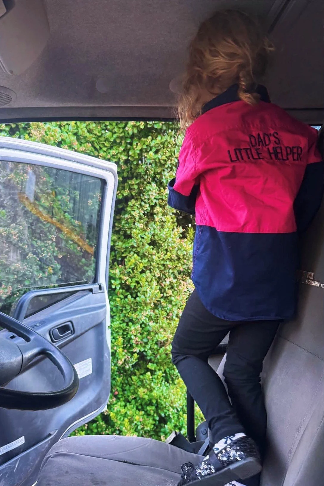 Child wearing a pink and navy jacket that reads 'Dad's Little Helper' standing on a seat inside a vehicle, looking outside through the open door with a bush visible outside.