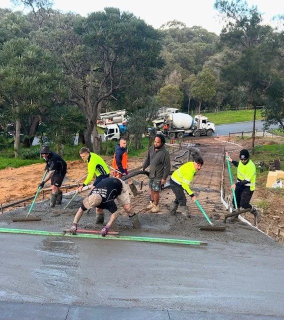 Construction workers smoothing and leveling wet concrete on a driveway or sidewalk, with construction vehicles and trees in the background.