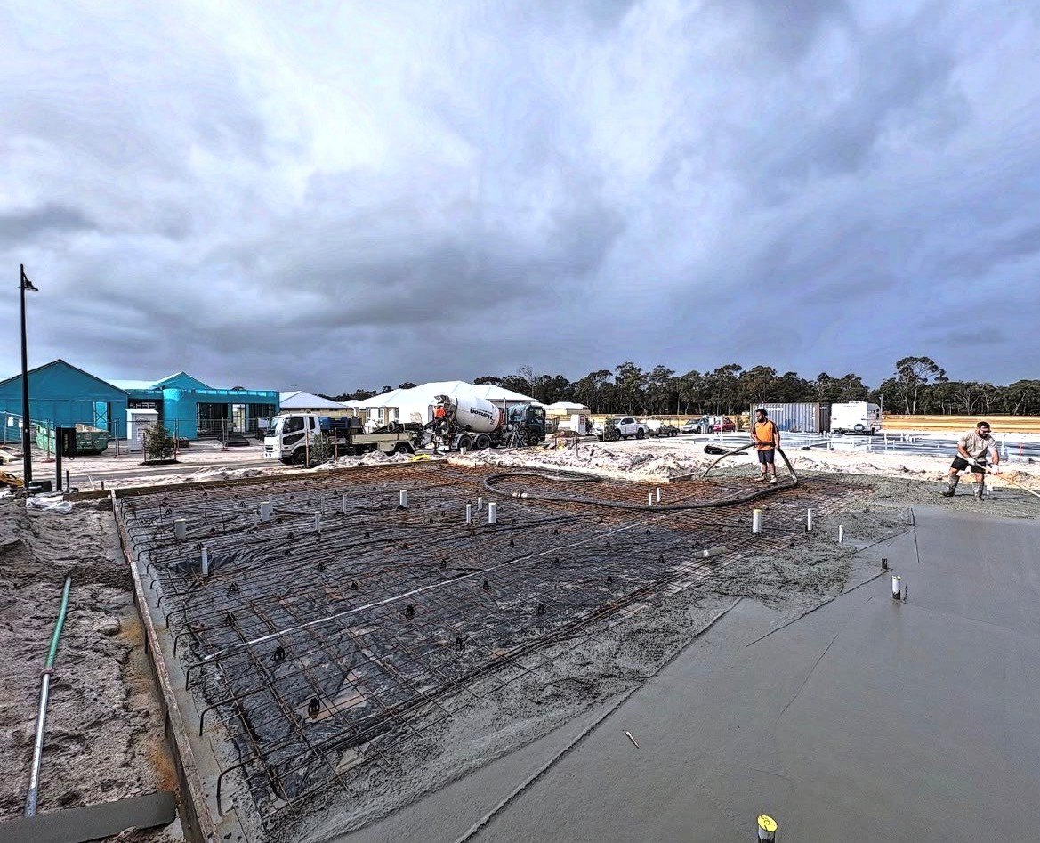 Construction site with workers pouring concrete, blue buildings, construction trucks, and cloudy sky.