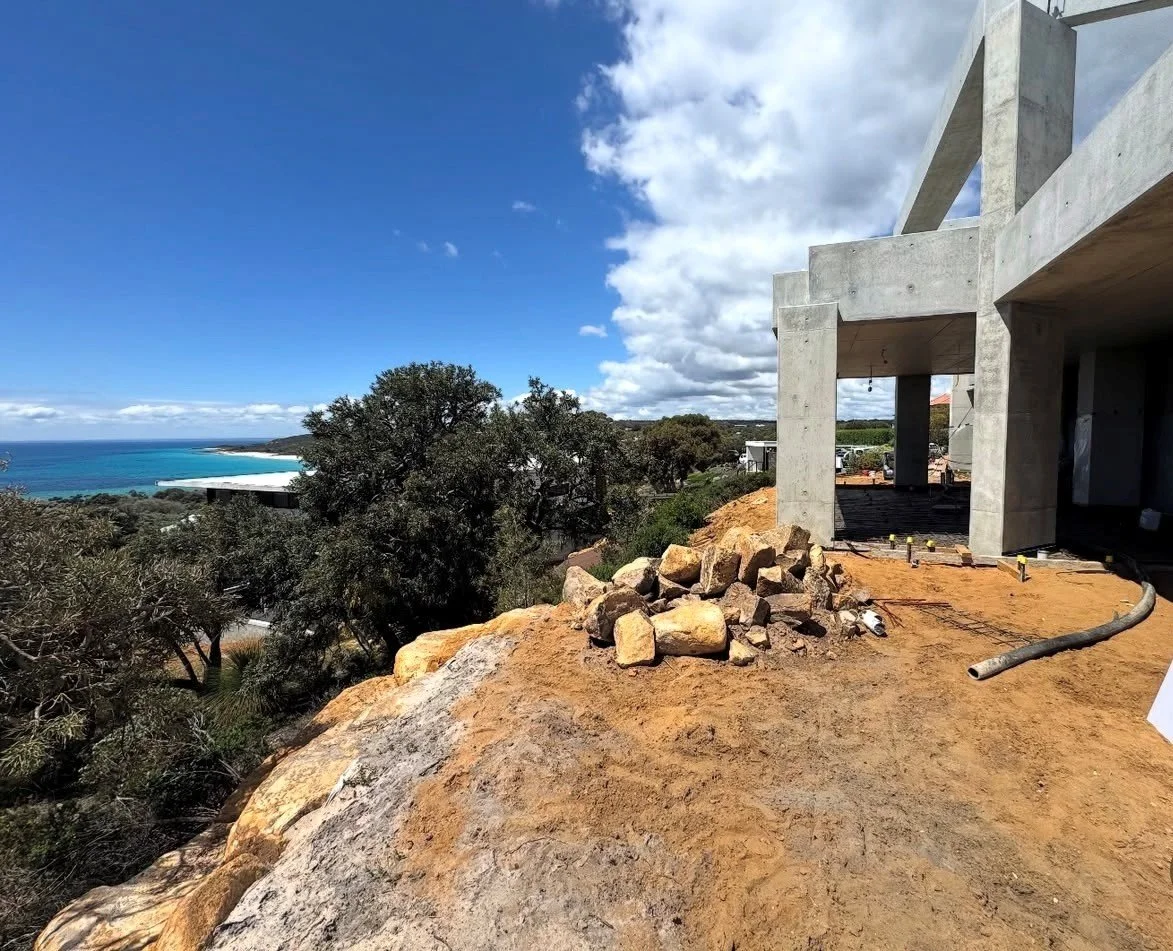 Construction site on a hill with a view of the ocean, showing a partially built concrete structure, rocks, and construction materials.