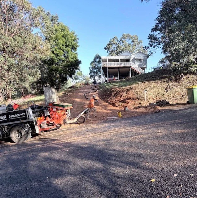 Construction workers paving a residential street, with a house on a hill in the background and trees surrounding the area.
