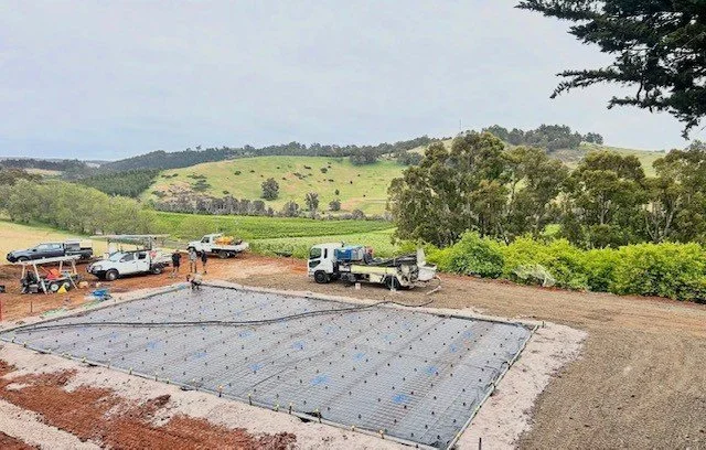 Construction site with a foundation prepared for pouring concrete, surrounded by trucks and workers, situated in a hilly rural area with trees and grass.