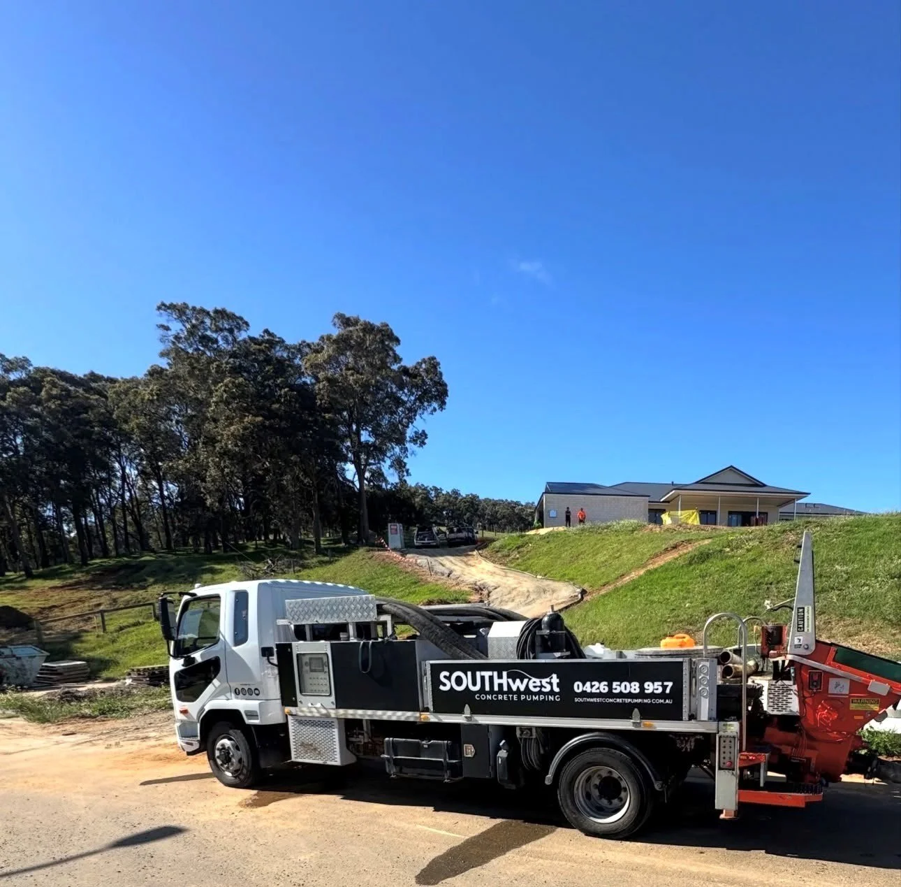 A concrete pumping truck parked on a construction site with a house and trees in the background under a blue sky.