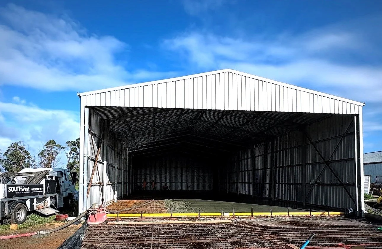 A large, open metal warehouse with a white roof under a partly cloudy blue sky, with construction equipment and materials nearby.