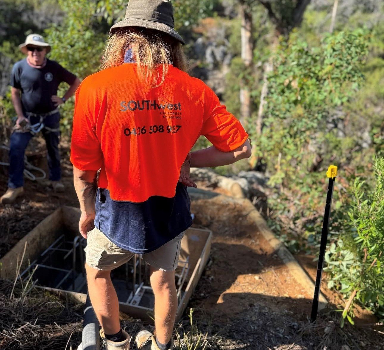 A person with long blonde hair wearing a black hat and an orange shirt from SOUTHwest Concrete Pumping is operating a piece of equipment outdoors in a wooded area, with another person standing behind them.