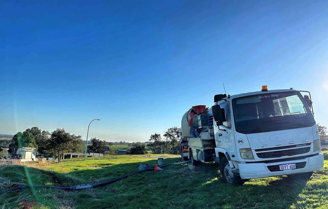 A white tanker truck parked on a grassy area, with pipes and equipment around it, under a clear blue sky.