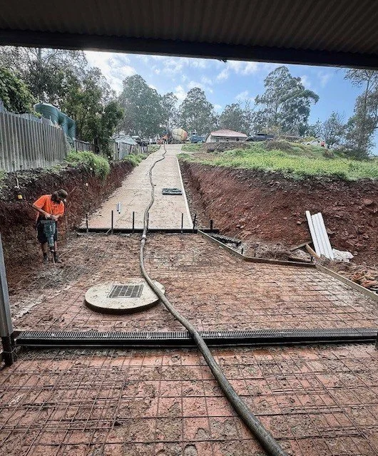 Worker pouring concrete on a leveled construction site outdoors, with a cloudy sky and trees in the background.