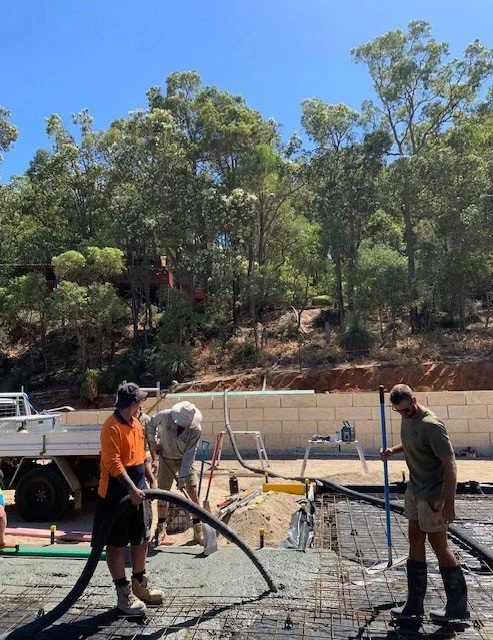 Three construction workers spray concrete onto a rebar framework at a construction site with trees in the background.