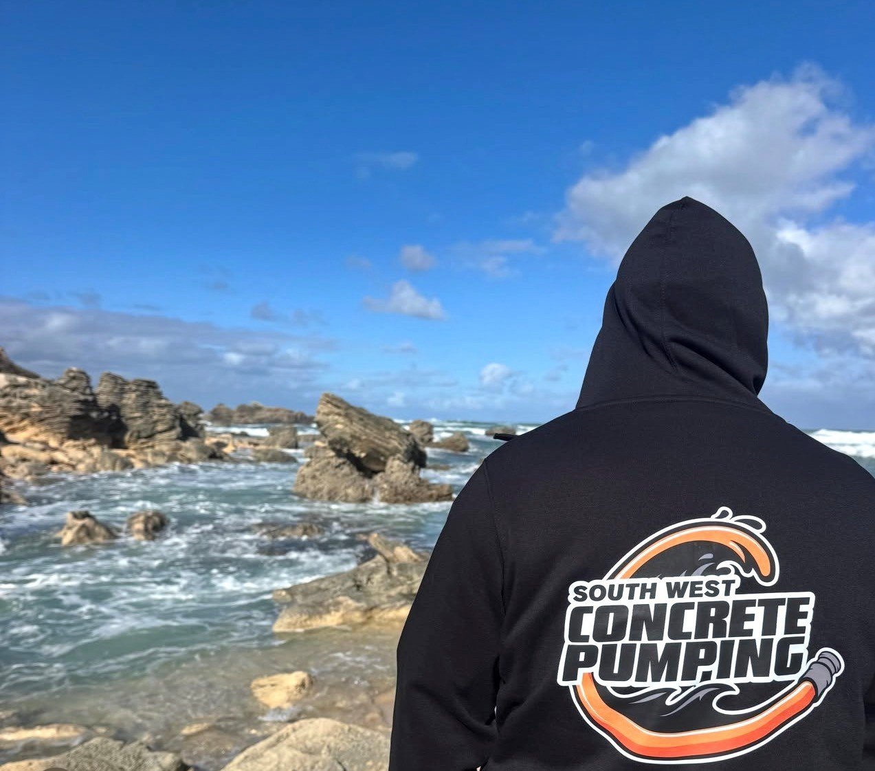 A person wearing a black hoodie with the logo 'South West Concrete Pumping' on the back, standing on a rocky shoreline with ocean waves and rocks, under a partly cloudy blue sky.