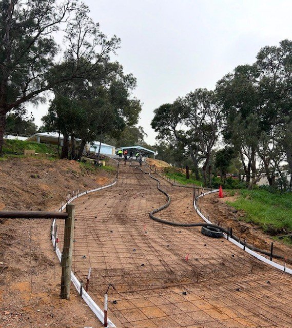 A construction site of a winding pathway under development with exposed rebar and construction hoses, surrounded by trees and grass, with some workers and equipment visible in the background.