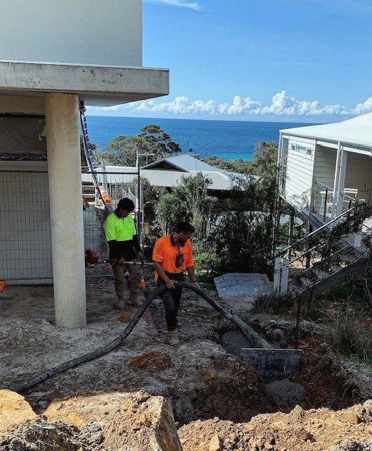Workers in bright safety shirts working on construction site near residential houses with ocean view in background.