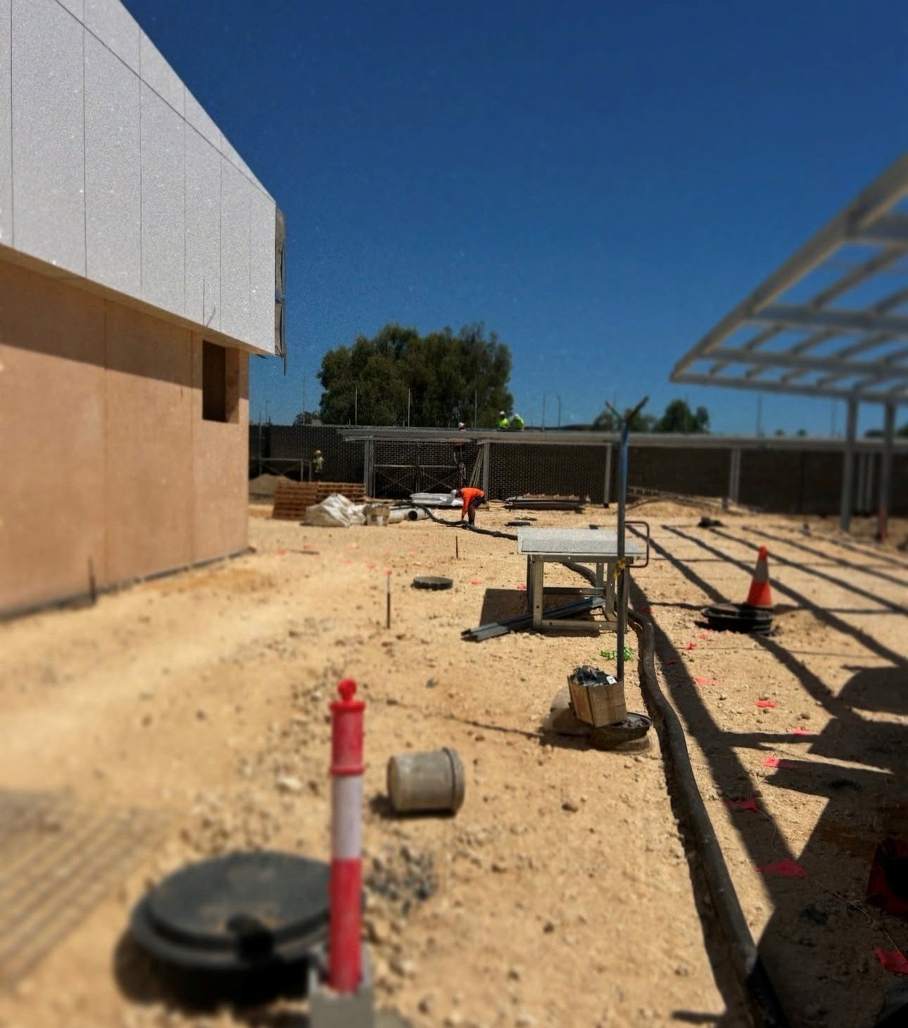 Construction site with workers, building materials, orange cones, and equipment under a clear blue sky.