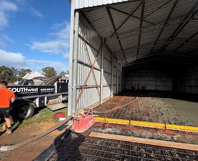 Construction workers pouring concrete inside a large metal warehouse with concrete pump equipment nearby.