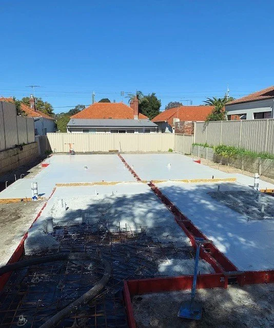 Residential backyard with a concrete foundation under construction, marked with red framing, surrounded by a wooden fence and neighboring houses with red roofs under a clear blue sky.