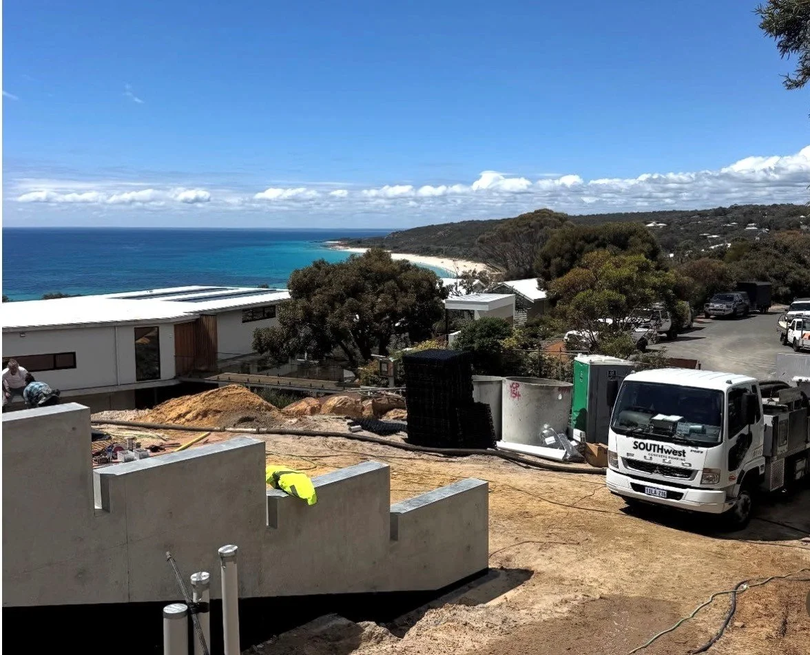 Construction site with trucks and equipment overlooking a beach and forested coastline under a blue sky with clouds.