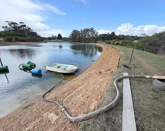 A shoreline with a partially submerged boat and floating barrels, with a hose running from the water to the land, under a partly cloudy sky.