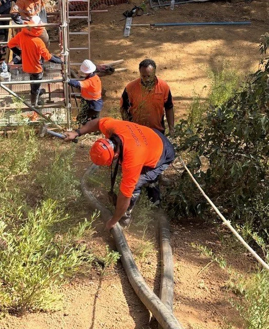Workers in orange shirts and helmets are installing or repairing large pipes in a landscaped outdoor area with bushes and dirt, using scaffolding for support.