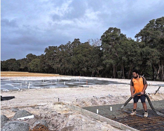 A construction worker in an orange safety vest and shorts working on a concrete foundation at a construction site with trees and cloudy sky in the background.