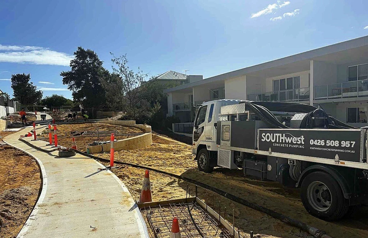 Construction workers working on a new sidewalk in a residential area with apartment buildings, a construction truck, traffic cones, and equipment under a partly cloudy sky.