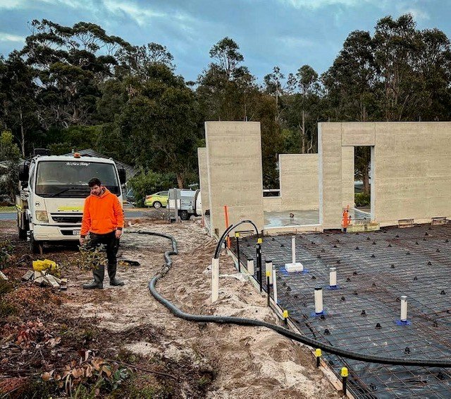 A construction worker in an orange jacket standing on a muddy construction site with concrete walls and a partially built floor, with trees in the background.