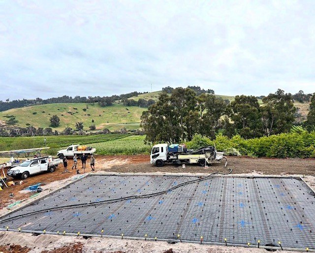 Construction site with a foundation and steel reinforcements, trucks, and construction workers, with a rural landscape of rolling hills and trees in the background.