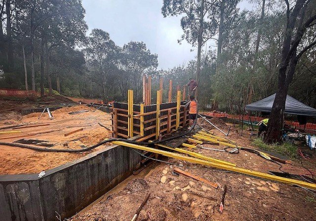 Construction site with workers building a concrete structure, surrounded by trees and construction equipment.