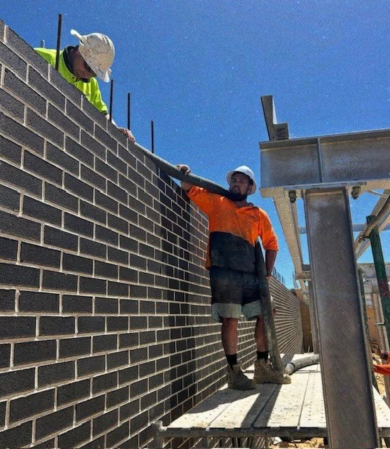 Two construction workers standing on scaffolding. One is leaning on the brick wall, and the other is looking down at the camera, both wearing safety helmets and work clothes. It is daytime with a clear sky.