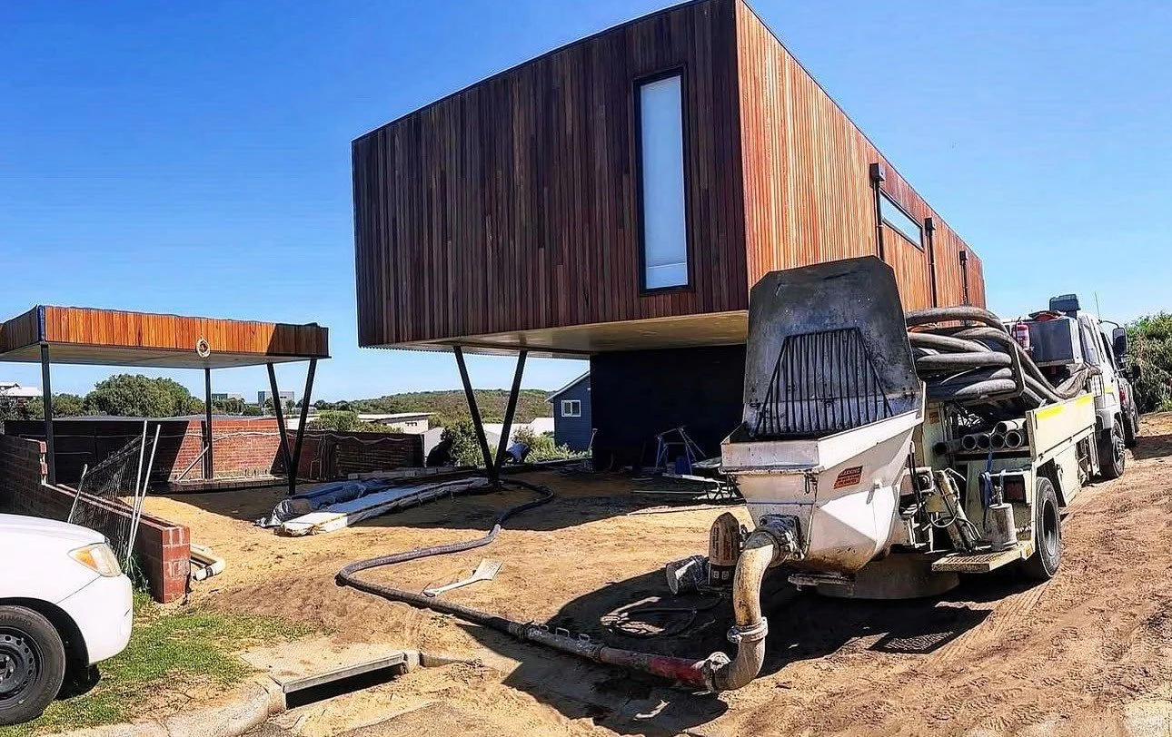 A modern wooden house built on stilts on a construction site with construction equipment and a white truck nearby.