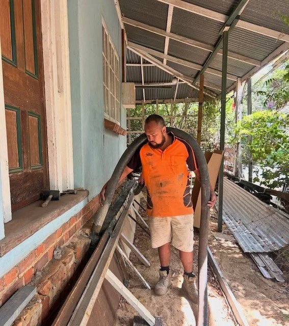Man in orange and black shirt standing outside a house, holding large black hoses, on a construction or renovation site with tools and building materials around.