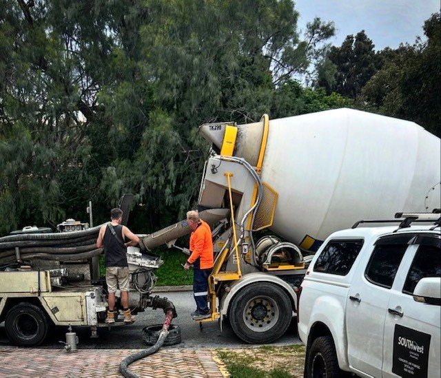 Two workers are pouring concrete from a mixer truck into a machine, with a white SUV parked nearby on a paved area with trees in the background.