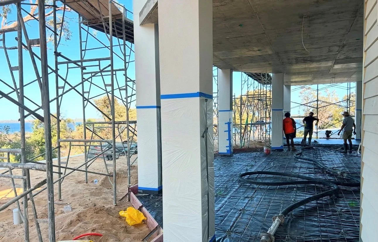 Construction workers on a building's underside, installing or working on the concrete floor, with scaffolding and scaffolding materials around them, and piping on the ground.