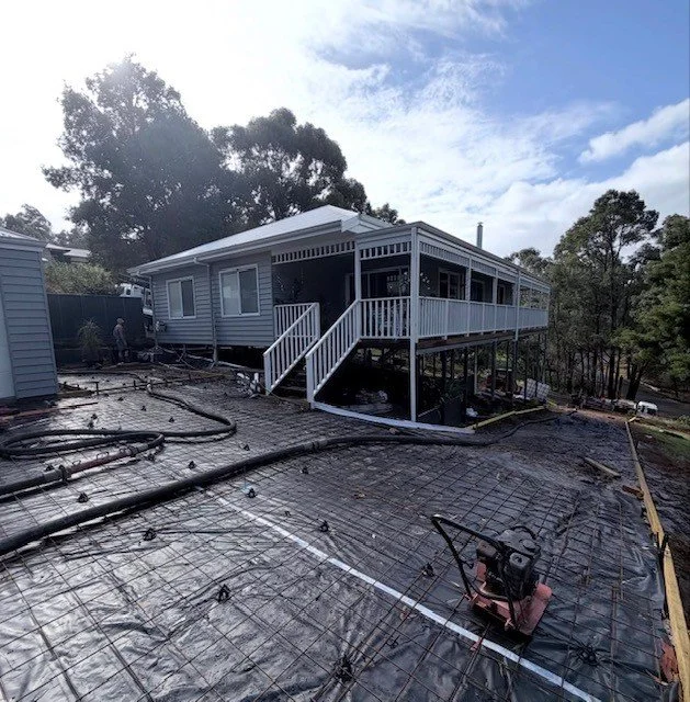 A house with a large elevated porch under construction, visible stairs, surrounded by black construction plastic, hoses, and rebar on the ground, with trees and a partly cloudy sky in the background.