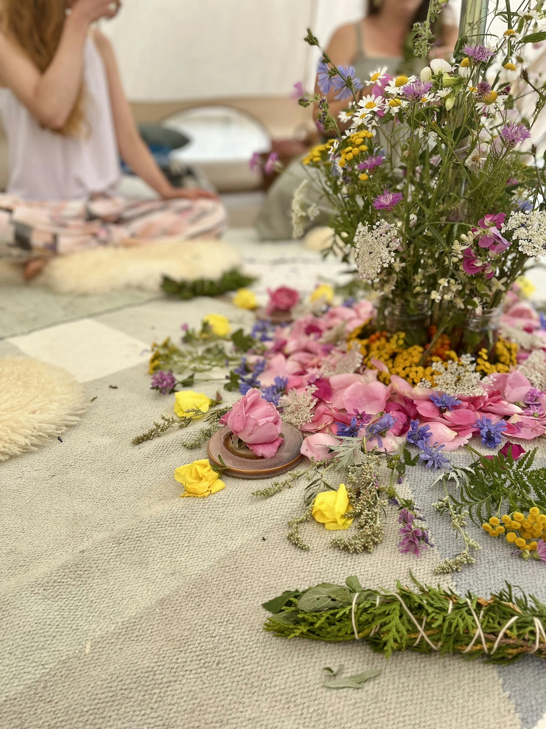 A large bouquet of colorful wildflowers including daisies, cornflowers, and other varieties sits on a textured beige floor, with loose flower petals scattered around. In the background, three women are seated on the floor, engaging in a flower arranging activity, with a blurred cozy home interior behind them.