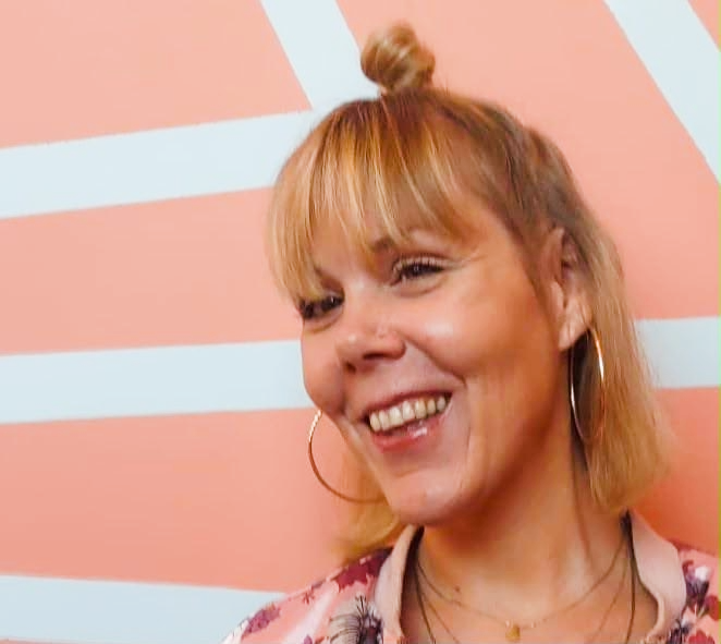 A woman with blonde hair styled in a small top bun, smiling and wearing large hoop earrings, standing in front of a pink and white background.