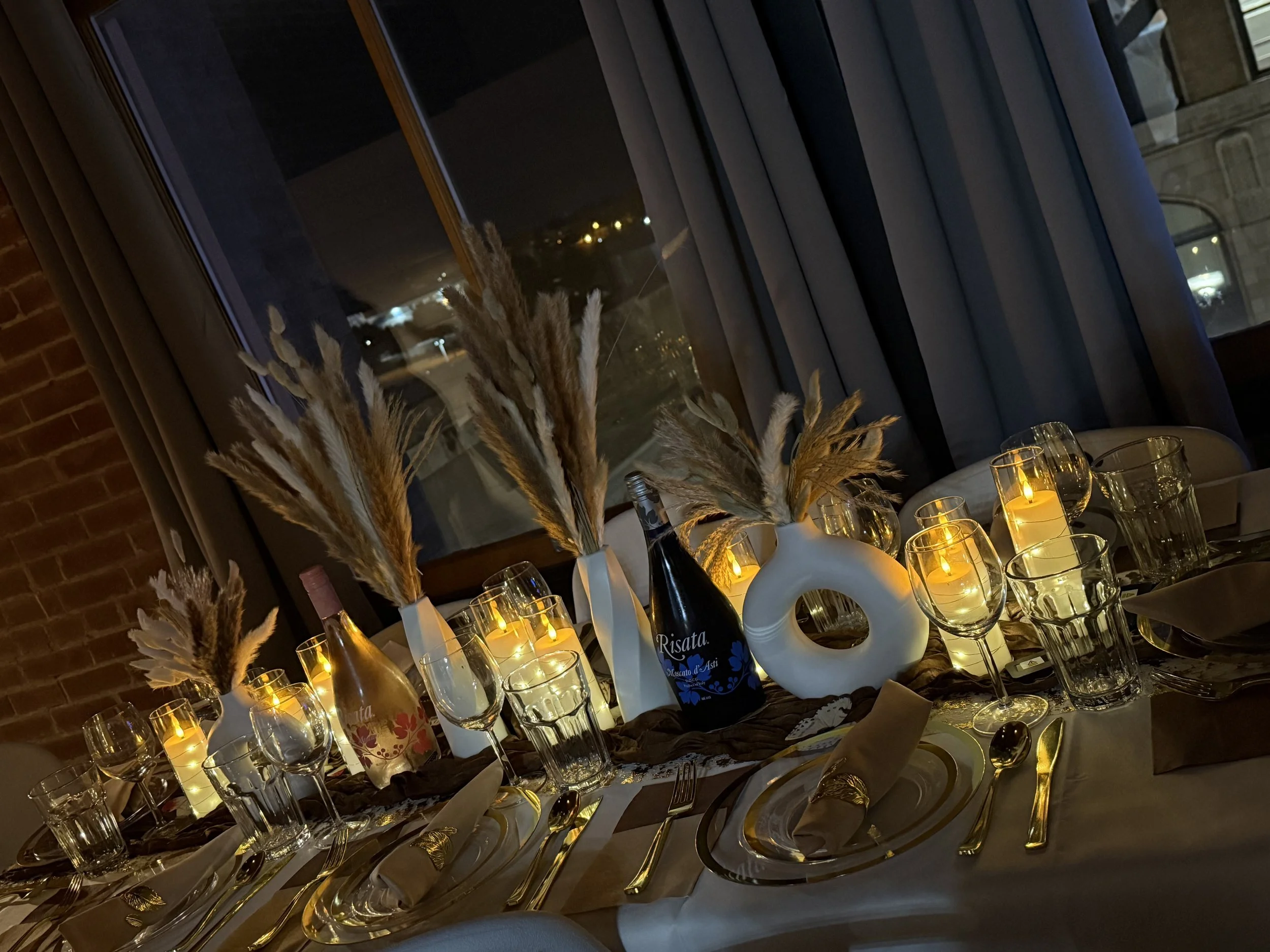 A dining table set with multiple wine glasses, candles, and white vases with dried pampas grass. There is a window in the background with a night view, including city lights and buildings. The setting appears elegant and cozy, suitable for a formal dinner.