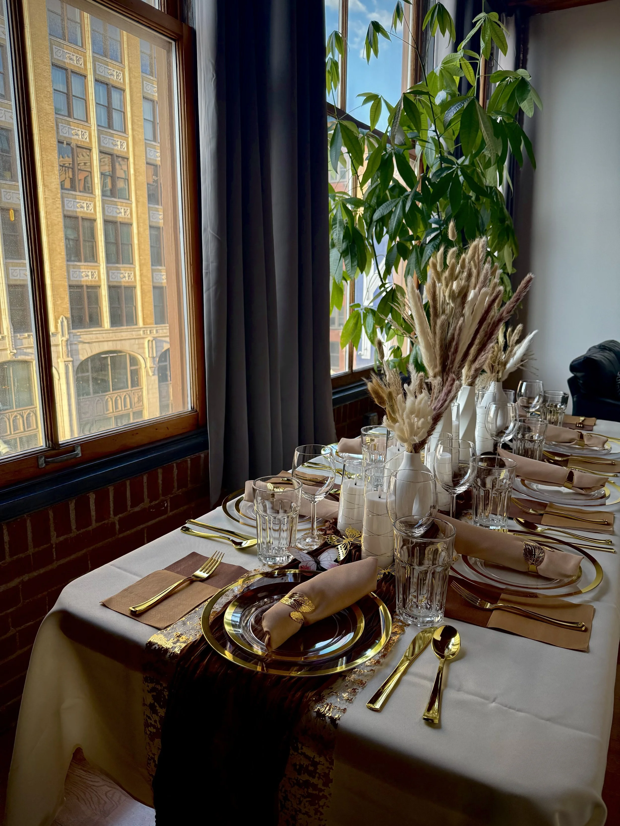 A formal dining table set with gold-rimmed plates, gold flatware, beige napkins with gold rings, empty glasses, and a centerpiece of dried pampas grass in a white vase. The room has a large window with black curtains, a brick wall, and a green plant.