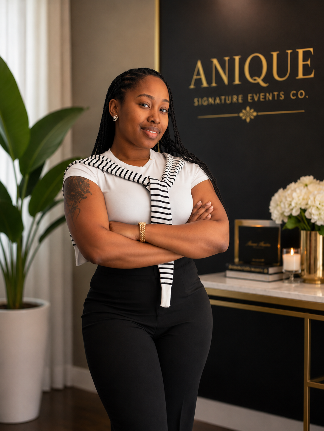 A woman standing with arms crossed in front of a black sign that reads 'ANIQUE SIGNATURE EVENTS CO.' in gold lettering, with a nearby table holding books, a candle, and flowers, in a well-decorated interior.