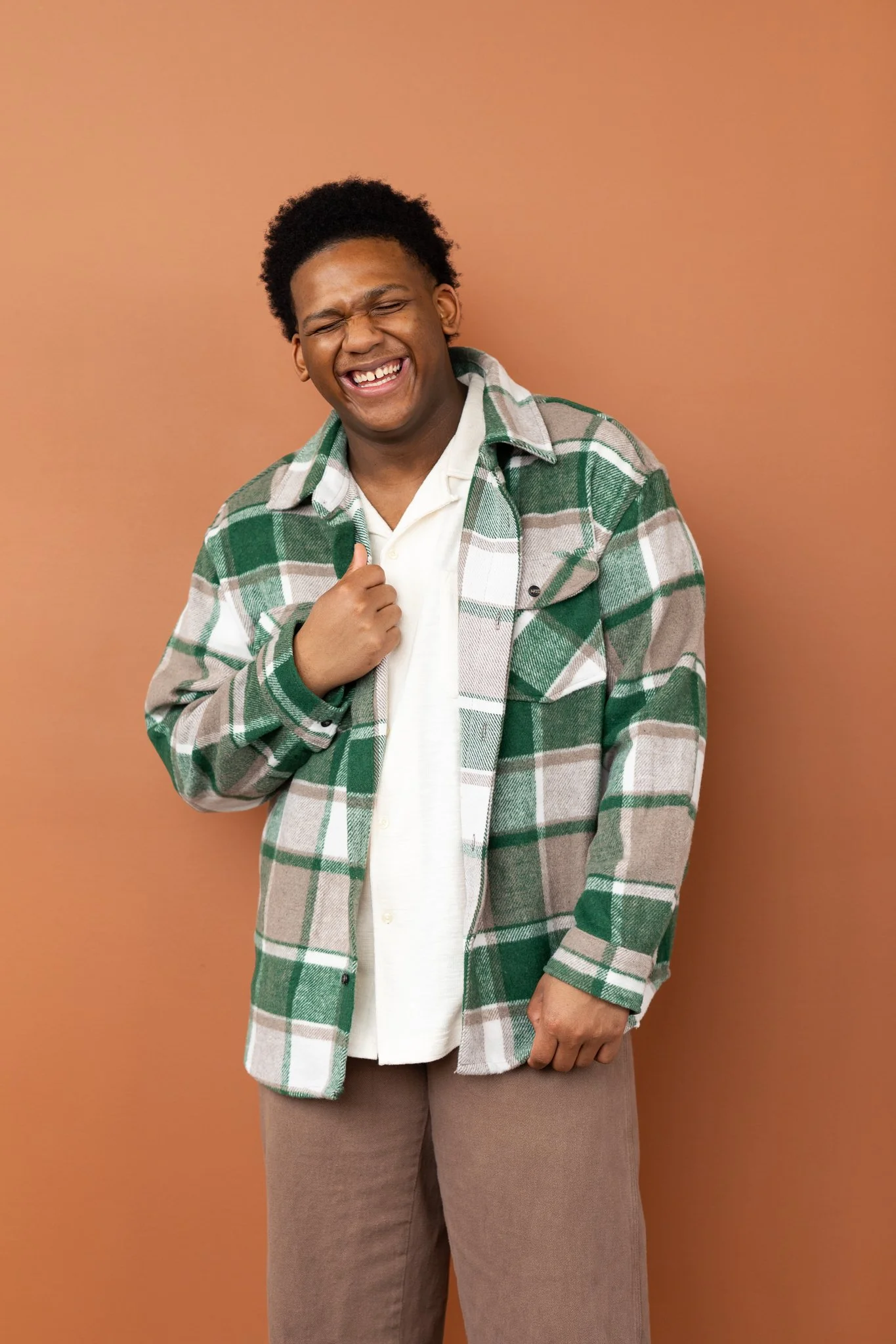 A person with dark skin, curly hair, laughing, wearing a green, white, and beige plaid jacket, a white shirt, and brown pants, standing against a plain orange-brown background.