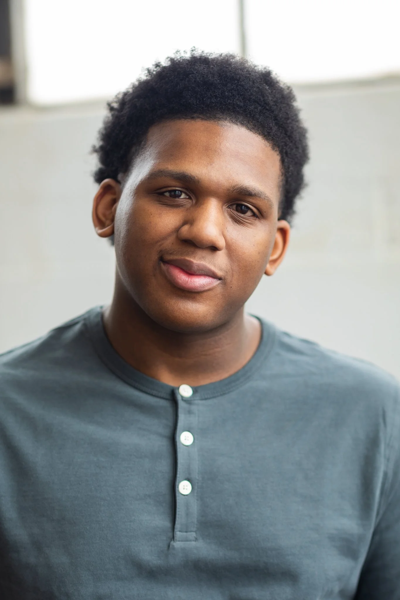 Close-up of a young man with dark skin and short, curly hair, wearing a dark gray henley shirt, looking at the camera with a slight smile.