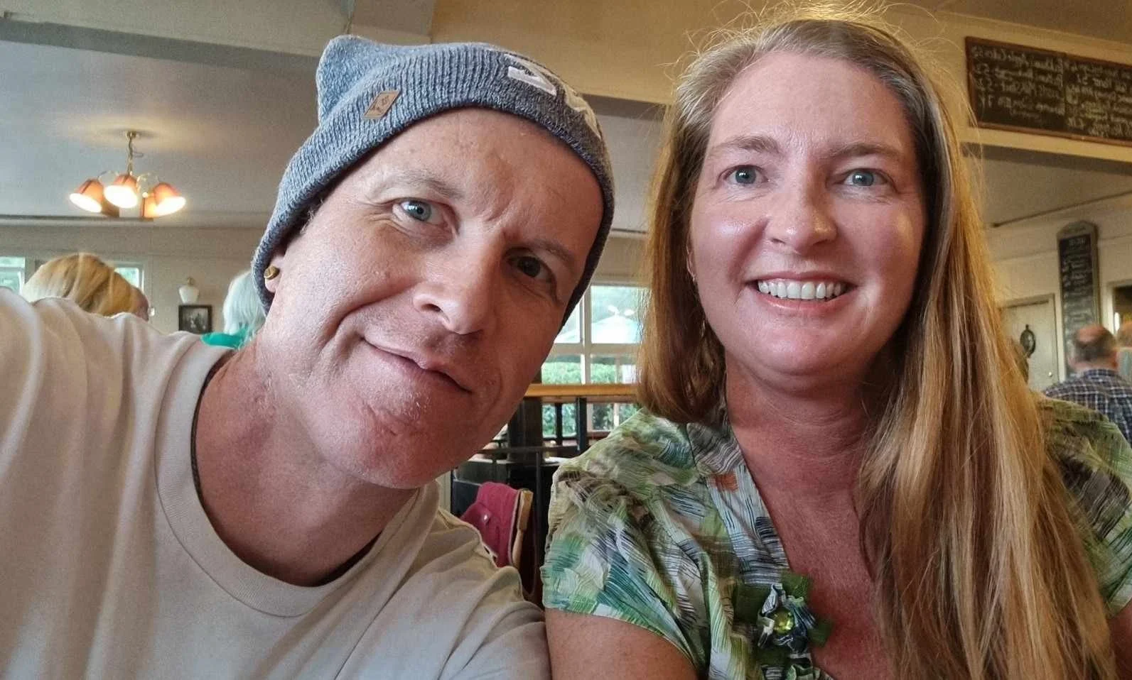 A man and woman taking a selfie inside a restaurant. The man is wearing a gray beanie and a light-colored shirt, while the woman has long reddish hair and is wearing a green patterned top. They are smiling and sitting close together, with other patrons and restaurant decor visible in the background.