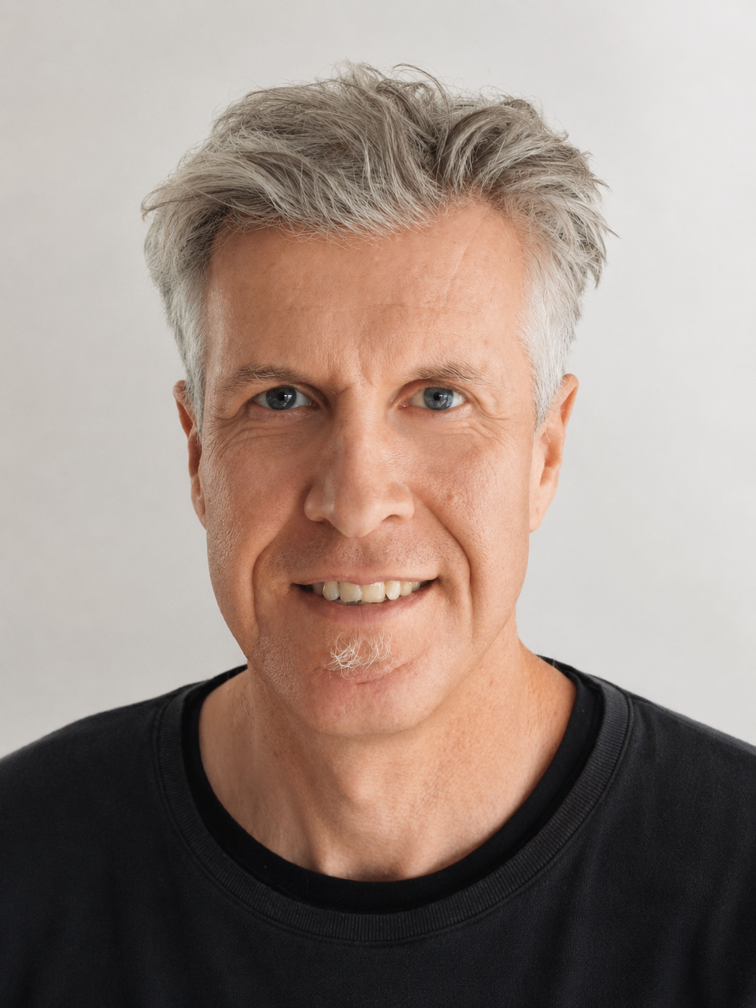 Close-up of a smiling middle-aged man with gray hair, blue eyes, and a small goatee, wearing a black shirt against a plain light background.
