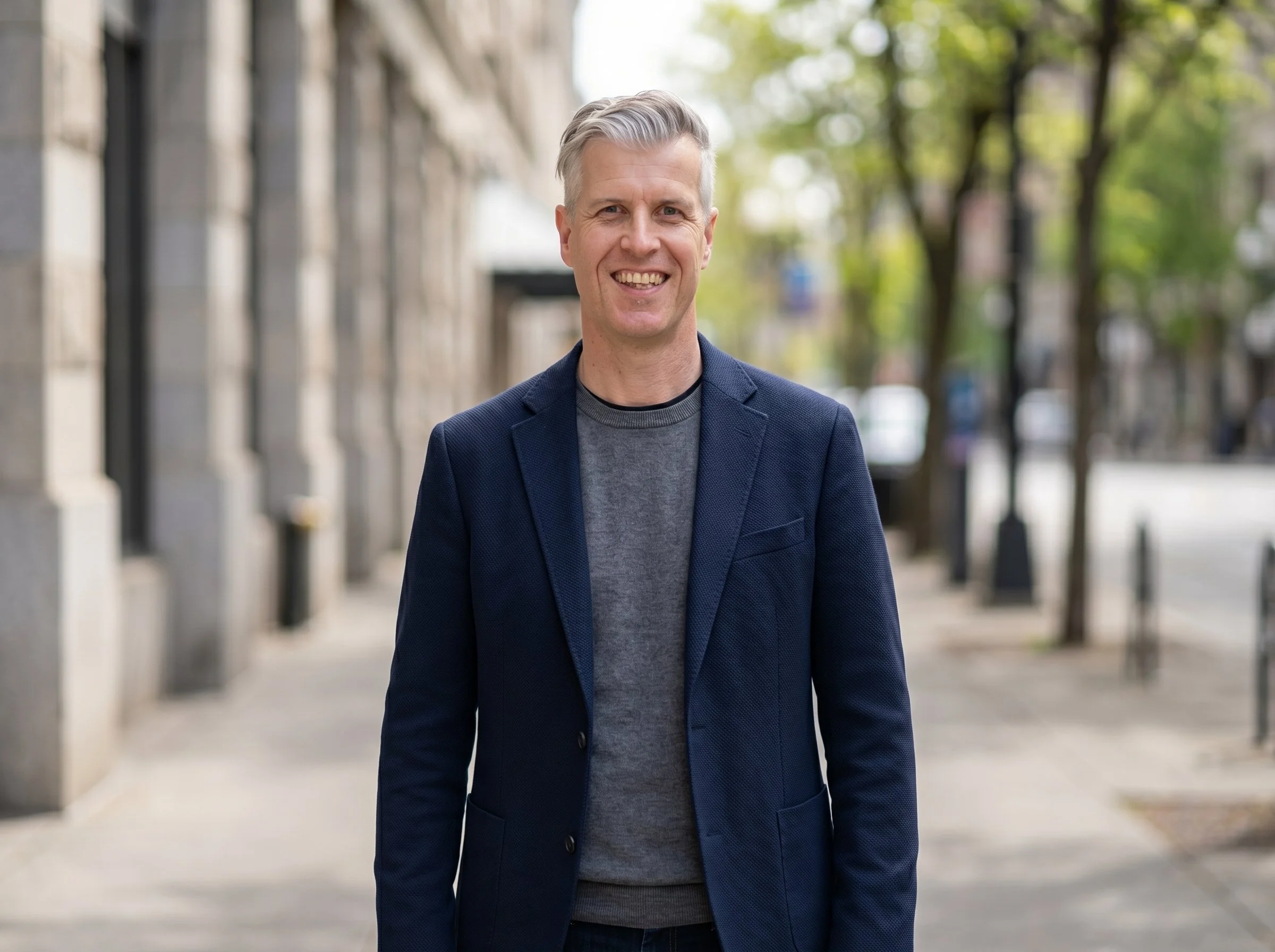 A smiling man with gray hair wearing a dark blazer and gray shirt walking outdoors on a city street with buildings and green trees in the background.