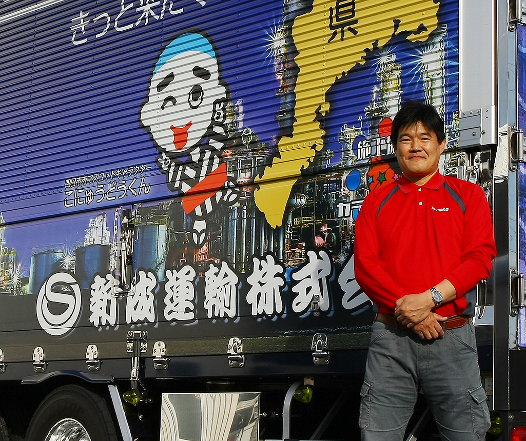 男性が赤いジャージを着て立っている。背景には鮮やかなイラストと日本語の文字が書かれたトラックがある。