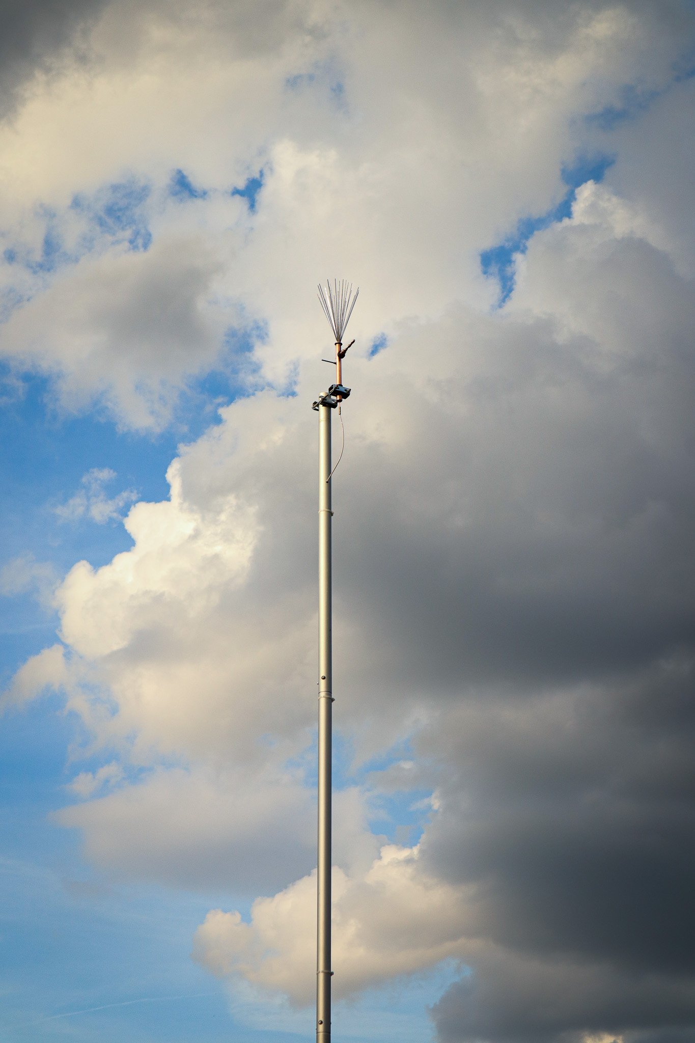 A tall vertical communication tower with a series of metal rods at the top, set against a cloudy sky during daytime.