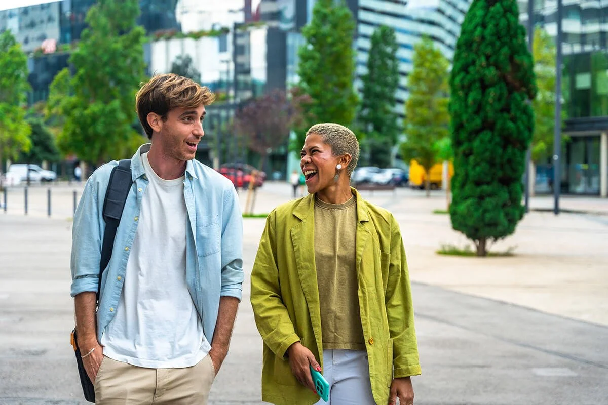 A man and woman walking and talking outdoors in an urban area with trees and modern buildings in the background, smiling and laughing.